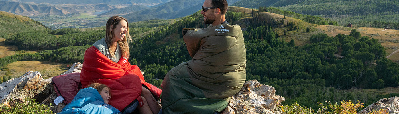 Two parents and a child sit atop a mountain overlook while wearing TETON Sports Celsius Sleeping Bags.
