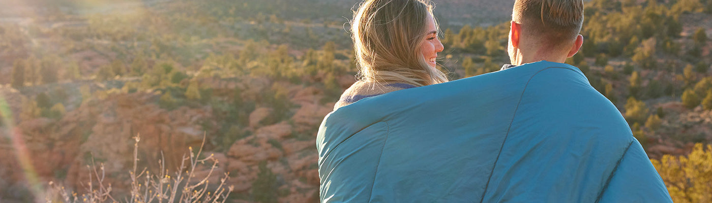 A man and a women sit together beneath a TETON Sports Outdoor Blanket watching the sunset.