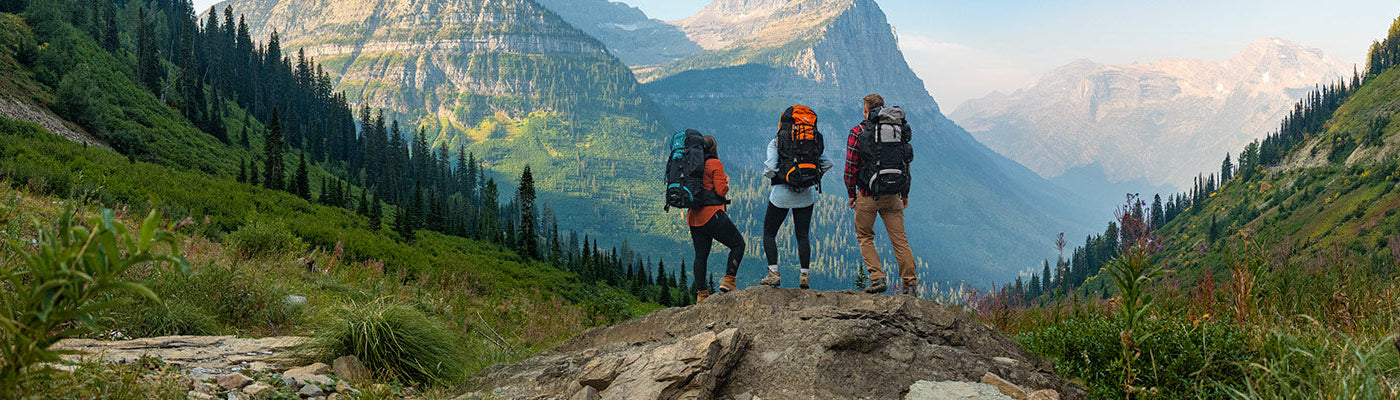 Three friends stands together atop a rocky outcrop wearing their TETON Sports Explorer & Scout backpacks, viewing a large mountain.