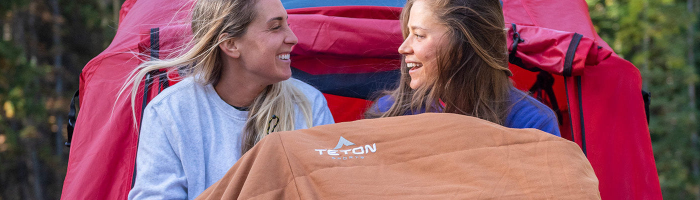 Two women snuggle together in their TETON Sports Canvas Mammoth Sleeping Bag on a cold morning.