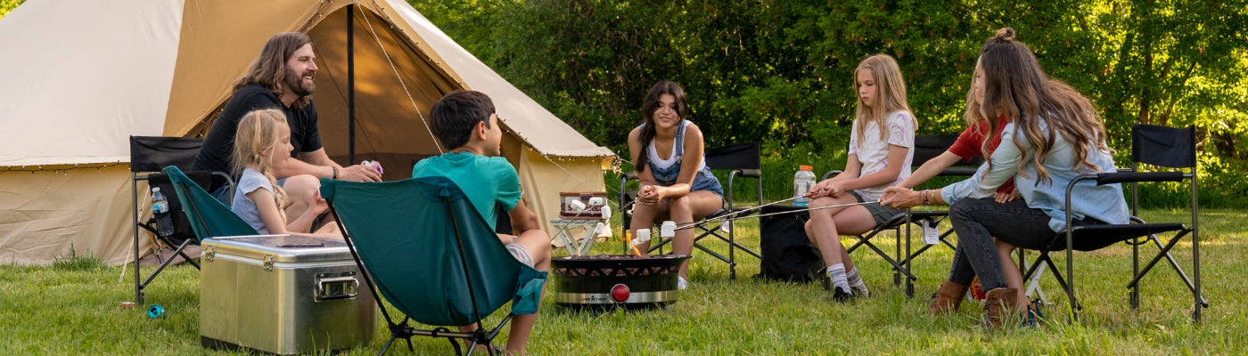 A family camps together around a campfire while their TETON Sports Sierra Canvas Bell Tent sits in the background.