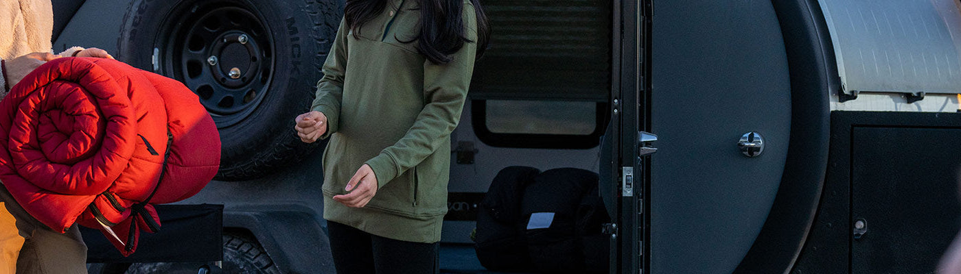 A man hands his rolled up sleeping bag to a woman with a camper in the background.