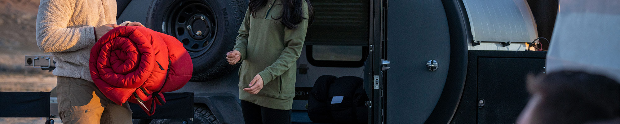 A man hands his rolled up sleeping bag to a woman with a camper in the background.