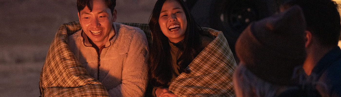 A couple laughs together by a campfire while snuggled up in a TETON Sports Evergreen Mammoth Sleeping Bag.