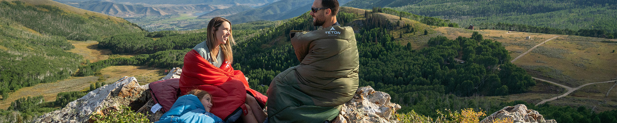 Two parents and a child sit atop a mountain overlook while wearing TETON Sports Celsius Sleeping Bags.