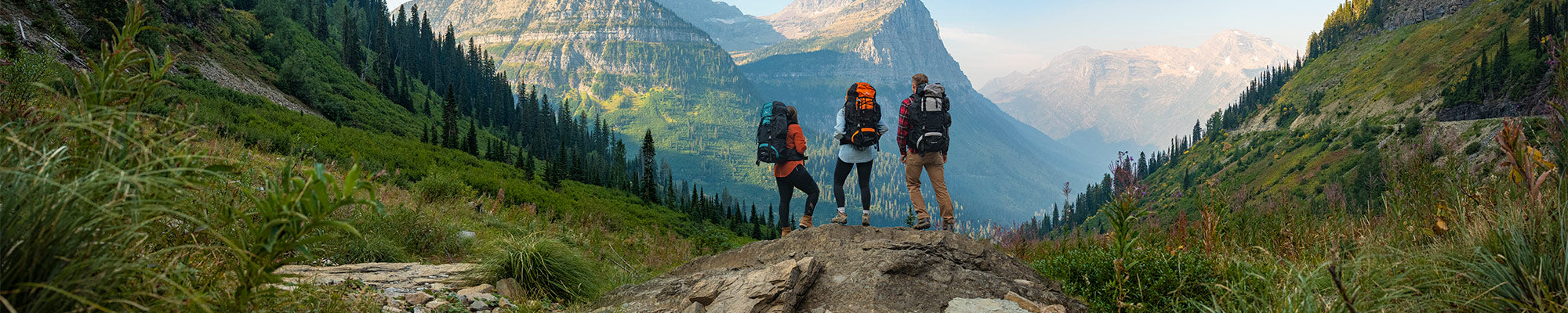 Three friends stands together atop a rocky outcrop wearing their TETON Sports Explorer & Scout backpacks, viewing a large mountain.