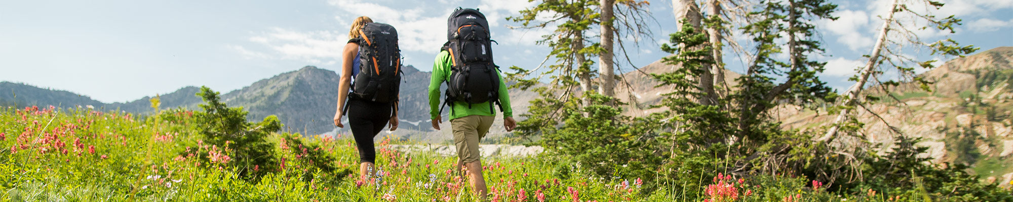 A couple backpacks together through a field of wildflowers while wearing TETON Sports backpacks.