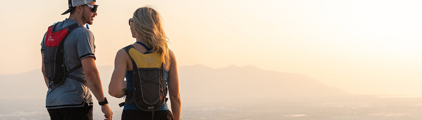 Two runners stop to sip from their TETON Sports TrailRunner 2L Hydration Packs while viewing a city from above.